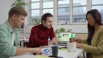 Wall Mural - Medium shot of designer presenting website interface templates for different devices on laptop screen to young colleagues taking notes sitting at desk in office