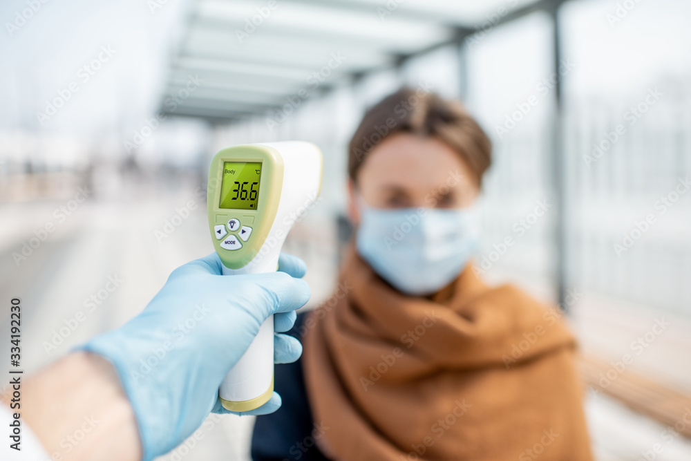 Measuring temperature with infrared thermometer of a young woman in face mask at a checkpoint during an epidemic outdoors. Concept of prevention the spread of the virus - obrazy, fototapety, plakaty 