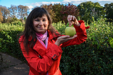 Beautiful smiling brunette girl in red coat holding fresh green apple on palm of her hand over nature background. Food, fruit and healthy nutrition