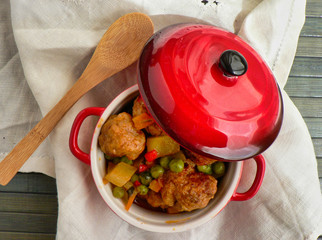 Canvas Print - Meatballs with potatoes and peas in a red casserole, wooden spoon and white tablecloth