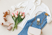 Blue Jeans, White Shirt, Heeled Sandals, Small Bag With Chain Strap, Accessories, Pink Tulips Flowers On Beige Background. Women's Stylish Spring Summer Outfit. Trendy Clothes. Flat Lay, Top View.