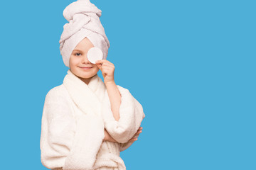 Smiling beautiful kid girl with towel on head hold cotton pad disk cleansing face skin with cleanser. Happy girl remove makeup enjoy healthy clean skincare beauty treatment concept