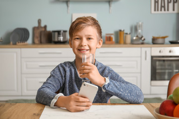 Wall Mural - Cute little boy with mobile phone drinking water in kitchen