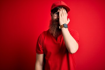 Canvas Print - Young handsome delivery man wearing glasses and red cap over isolated background covering one eye with hand, confident smile on face and surprise emotion.