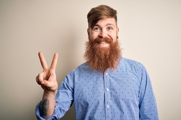 Sticker - Handsome Irish redhead business man with beard standing over isolated background smiling with happy face winking at the camera doing victory sign. Number two.