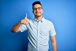 © Krakenimages.com - Young handsome man wearing casual summer shirt and glasses over isolated blue background doing happy thumbs up gesture with hand. Approving expression looking at the camera showing success.
