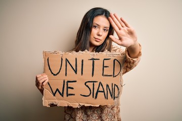 Wall Mural - Young beautiful brunette woman asking for union holding banner with unity message with open hand doing stop sign with serious and confident expression, defense gesture