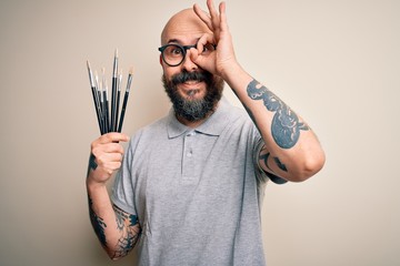 Wall Mural - Handsome bald artist man with beard and tattoo painting using painter brushes with happy face smiling doing ok sign with hand on eye looking through fingers