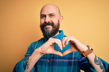 Sticker - Handsome bald man with beard wearing casual shirt standing over isolated yellow background smiling in love showing heart symbol and shape with hands. Romantic concept.