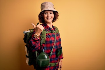 Poster - Middle age curly hair hiker woman hiking wearing backpack and water canteen using binoculars smiling with happy face looking and pointing to the side with thumb up.