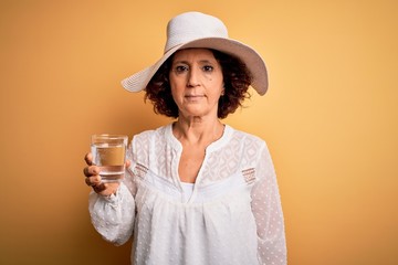 Poster - Middle age curly woman on vacation drinking glass of water over isolated yellow background with a confident expression on smart face thinking serious