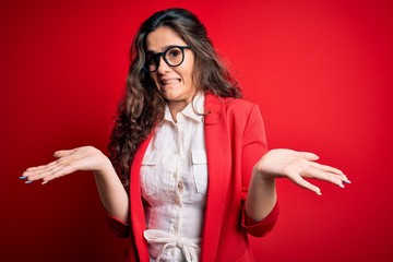 Poster - Young beautiful woman with curly hair wearing jacket and glasses over red background clueless and confused expression with arms and hands raised. Doubt concept.