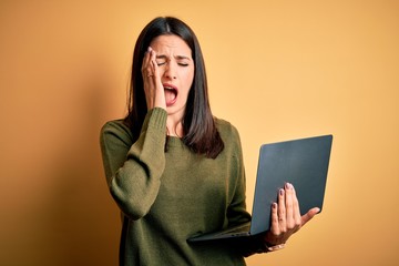 Young brunette woman with blue eyes working using computer laptop over yellow background Yawning tired covering half face, eye and mouth with hand. Face hurts in pain.