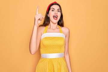 Canvas Print - Young beautiful pin up woman wearing 50s fashion vintage dress over yellow background pointing finger up with successful idea. Exited and happy. Number one.