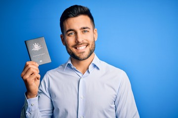 Young handsome tourist man holding australia australian passport id over blue background with a happy face standing and smiling with a confident smile showing teeth