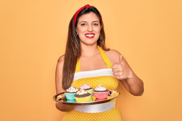 Young hispanic pin up woman wearing fashion sexy 50s style holding sweet cupcakes happy with big smile doing ok sign, thumb up with fingers, excellent sign