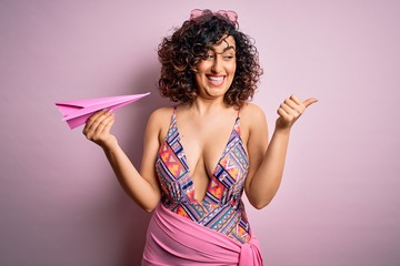 Wall Mural - Young beautiful curly arab woman on vacation wearing swimsuit holding pink paper airplane pointing and showing with thumb up to the side with happy face smiling