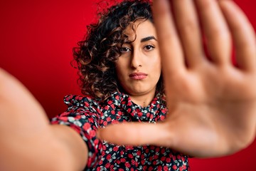 Sticker - Young beautiful curly arab woman wearing dress making selfie by camera over red background with open hand doing stop sign with serious and confident expression, defense gesture