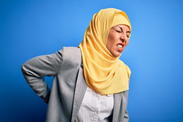 Wall Mural - Middle age brunette business woman wearing muslim traditional hijab over blue background Suffering of backache, touching back with hand, muscular pain