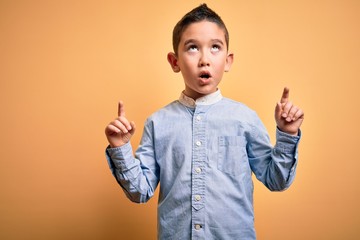 Wall Mural - Young little boy kid wearing elegant shirt standing over yellow isolated background amazed and surprised looking up and pointing with fingers and raised arms.