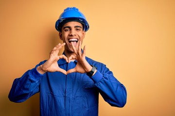 Poster - Young handsome african american worker man wearing blue uniform and security helmet smiling in love showing heart symbol and shape with hands. Romantic concept.