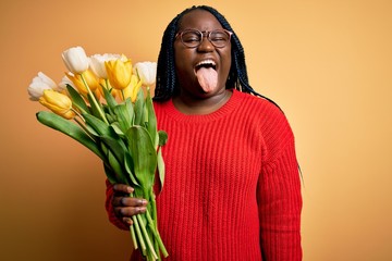 Sticker - Young african american plus size woman with braids holding bouquet of yellow tulips flower sticking tongue out happy with funny expression. Emotion concept.