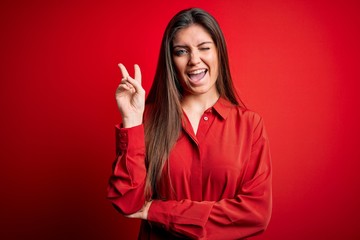 Young beautiful woman with blue eyes wearing casual shirt standing over red background smiling with happy face winking at the camera doing victory sign. Number two.