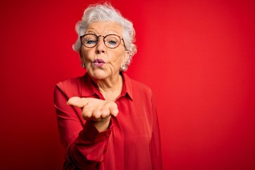 Wall Mural - Senior beautiful grey-haired woman wearing casual shirt and glasses over red background looking at the camera blowing a kiss with hand on air being lovely and sexy. Love expression.