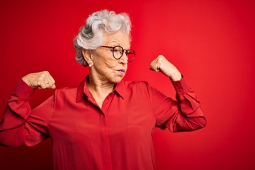 Senior beautiful grey-haired woman wearing casual shirt and glasses over red background showing arms muscles smiling proud. Fitness concept.