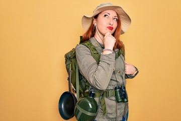 Wall Mural - Young redhead backpacker woman hiking wearing backpack and hat over yellow background with hand on chin thinking about question, pensive expression. Smiling and thoughtful face. Doubt concept.