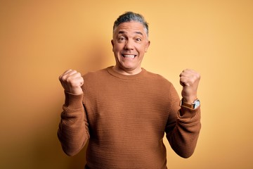 Poster - Middle age handsome grey-haired man wearing casual sweater over yellow background celebrating surprised and amazed for success with arms raised and open eyes. Winner concept.