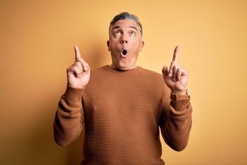 Poster - Middle age handsome grey-haired man wearing casual sweater over yellow background amazed and surprised looking up and pointing with fingers and raised arms.