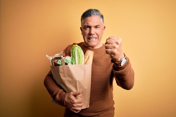 Poster - Middle age handsome grey-haired man holding paper bag with food over yellow background annoyed and frustrated shouting with anger, crazy and yelling with raised hand, anger concept