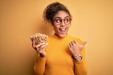 Wall Mural - Young african american afro girl holding bowl with healthy peanuts over yellow background pointing and showing with thumb up to the side with happy face smiling