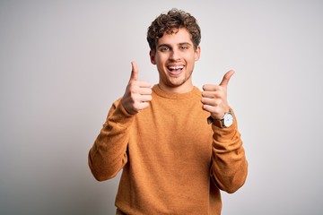 Young blond handsome man with curly hair wearing casual sweater over white background success sign doing positive gesture with hand, thumbs up smiling and happy. Cheerful expression and winner