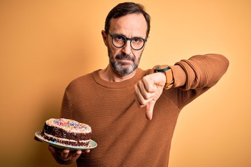 Sticker - Middle age hoary man holding birthday cake standing over isolated yellow background with angry face, negative sign showing dislike with thumbs down, rejection concept