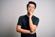 Young handsome chinese man wearing black t-shirt and glasses over white background looking confident at the camera with smile with crossed arms and hand raised on chin. Thinking positive.