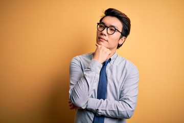 Young handsome chinese businessman wearing glasses and tie over yellow background with hand on chin thinking about question, pensive expression. Smiling with thoughtful face. Doubt concept.