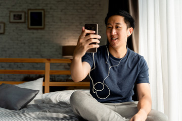 Portrait of happy 30s aged Asian man in casual clothing making facetime video calling with smartphone at home. He's waving at people on phone screen. Using conferencing meeting online app concept