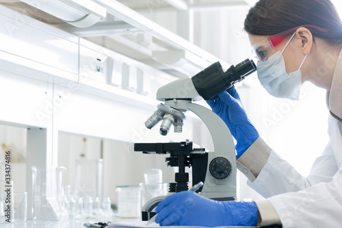 Fotomural Young woman in whitecoat, gloves, eyewear and mask bending over microscope