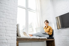 Young Woman Dressed Casually Working On Laptop While Sitting On The Window Sill At Home. Work From Home At Cozy Atmosphere Concept