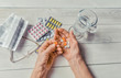 Senior hands with pills and drugs on table, glass of water. Wrinkled hands of old woman holding colorful tablets, glasses and prescription wooden background. Old age. Health care for the elderly.