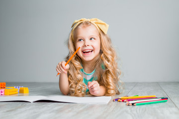 happy little child, a charming blonde baby is lying comfortably on the wooden floor, drawing on paper with colored pencils