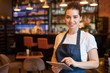 © pressmaster - Young smiling waitress in apron and t-shirt standing in front of camera