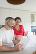 © Rafa Fernandez - Happy pregnant woman with her husband sitting at home. Smiling couple consulting a book at the kitchen.