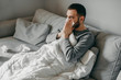© lesia17 - Young sick bearded european man sneezes into napkin at home on gray sofa with white blanket. Disease, protection, coronavirus, virus, disease, flu, respiratory dressing.