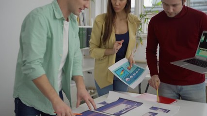 Wall Mural - Tilt down from above shot of young startup team of three specialists standing around table in office and discussing website interface templates presented on laptop and printed on paper
