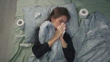 The Sick Girl Lies In Bed Under The Covers And Blows Her Nose. Toilet Paper In A Coronavirus Pandemic Like A Handkerchief. Coronavirus Protection. Home Treatment At The Time Of Carntin.
