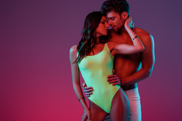 Wall Mural - handsome shirtless man hugging sexy girl in swimsuit touching his neck on purple background with gradient