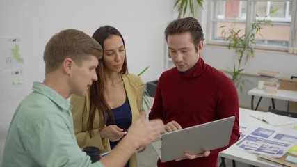 Wall Mural - Group of three young specialists, two men and woman, looking at laptop screen and discussing ideas standing in office
